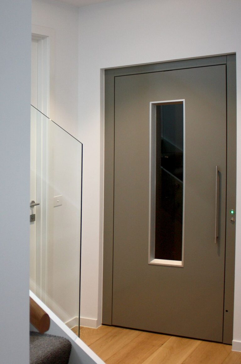 Grey ElfoLift door with a narrow vertical glass slot and a long handle beside a glass balustrade