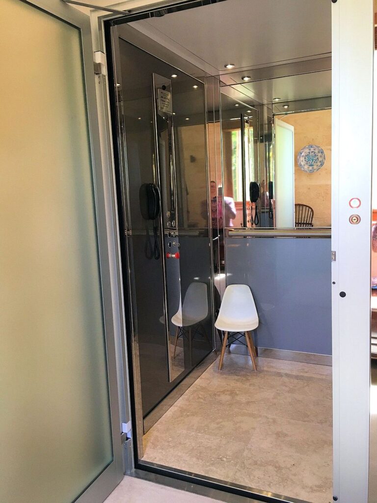 Open ElfoLift door showing the mirrored cabin, a white chair and a stone tile floor