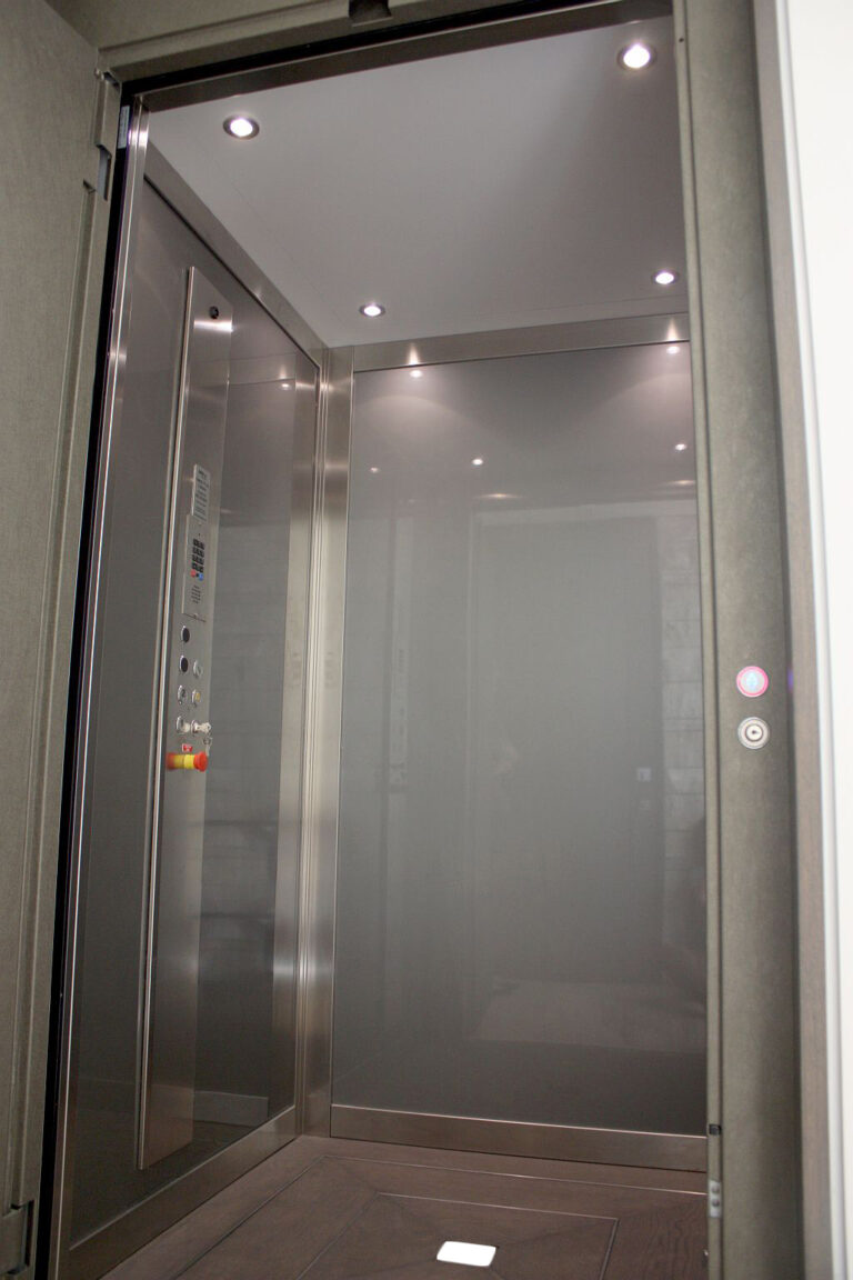 Inside the Laguna Beach lift cabin: grey skinplate walls, brushed stainless steel profiles, downlights, a side control panel and a timber floor with a stone centrepiece