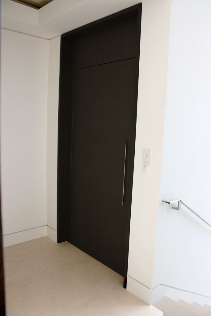 Tall dark lift landing door with a long steel handle in a white wall beside the stair handrail