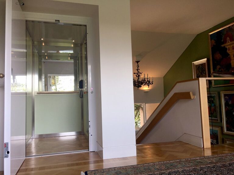 Glass fronted ElfoLift beside a timber handrail and stair, with a chandelier beyond, in an Auchenflower home