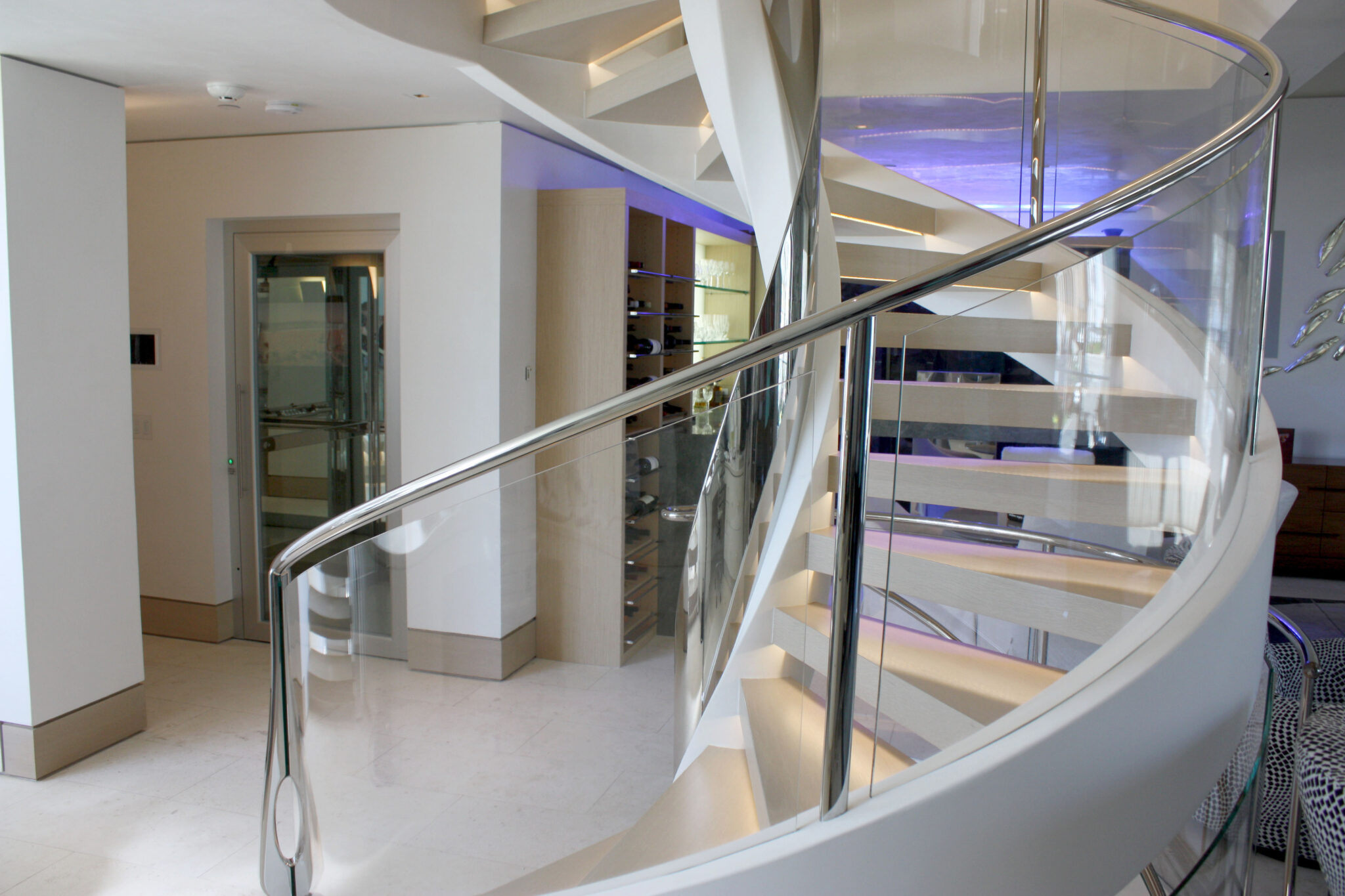 White spiral staircase with a glass balustrade beside the glass-doored home lift in a Californian beach house