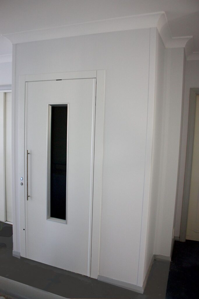 The finished white lift door with a narrow glass window set into the hallway wall