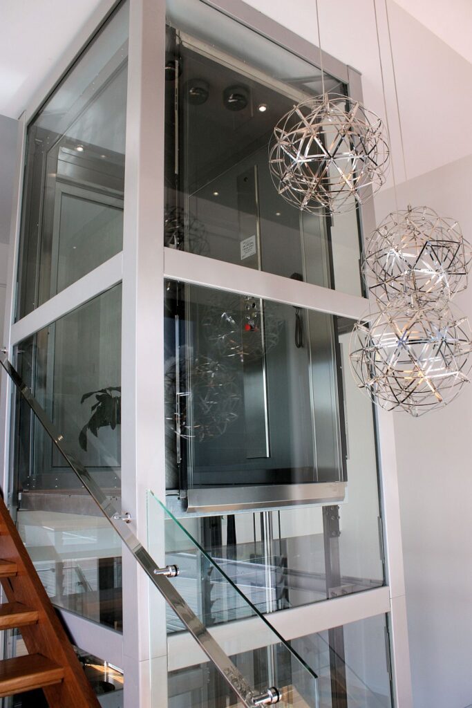Tall glass lift shaft with a stainless frame beside a staircase and pendant lights in an Ashgrove home