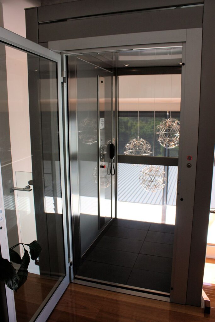 Open glass landing door showing the lift cabin with a dark tiled floor, stainless frame and pendant lights beyond