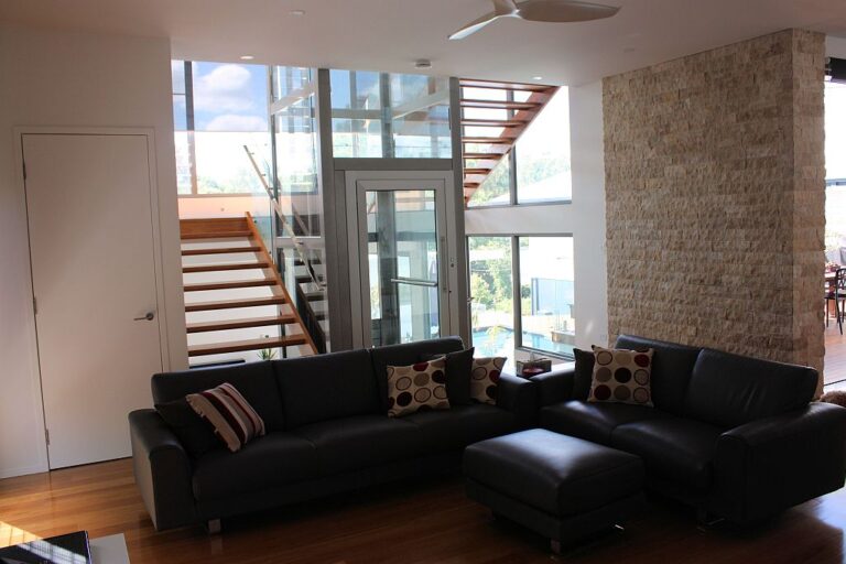 Living room with a black leather sofa and a stone feature wall, with the glass lift and open-tread staircase beyond