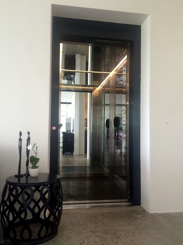Stainless steel lift with a mirrored cabin and LED strip behind a dark door frame, beside a black side table and sculpture