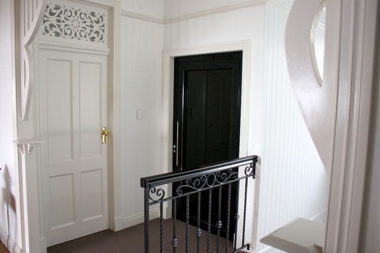 Dark lift door beside a white panelled door and ornate black iron balustrade on a landing