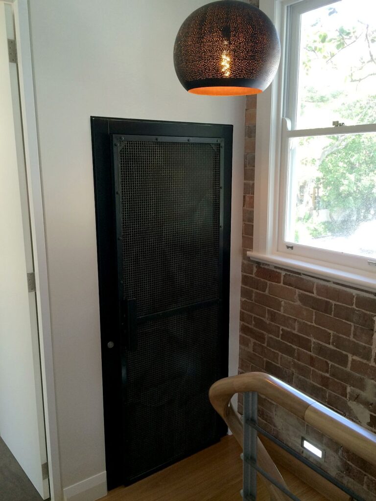 Black steel mesh landing door on the middle level beside an exposed brick wall and a woven pendant light