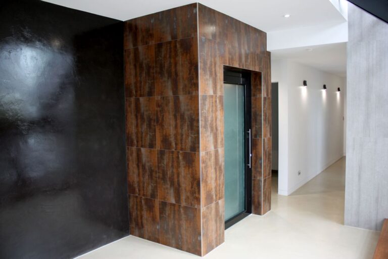 Rust coloured tile feature wall enclosing a lift with a frosted glass door beside black wall