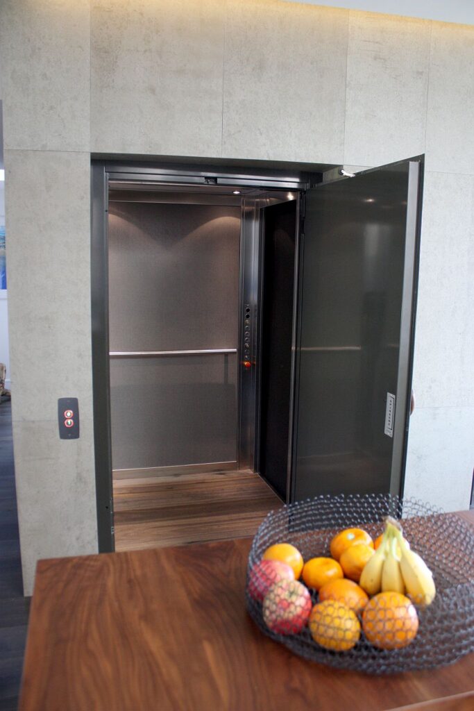 Open lift door in a stone-look enclosure showing the stainless steel cabin, beside a bowl of fruit on a timber bench