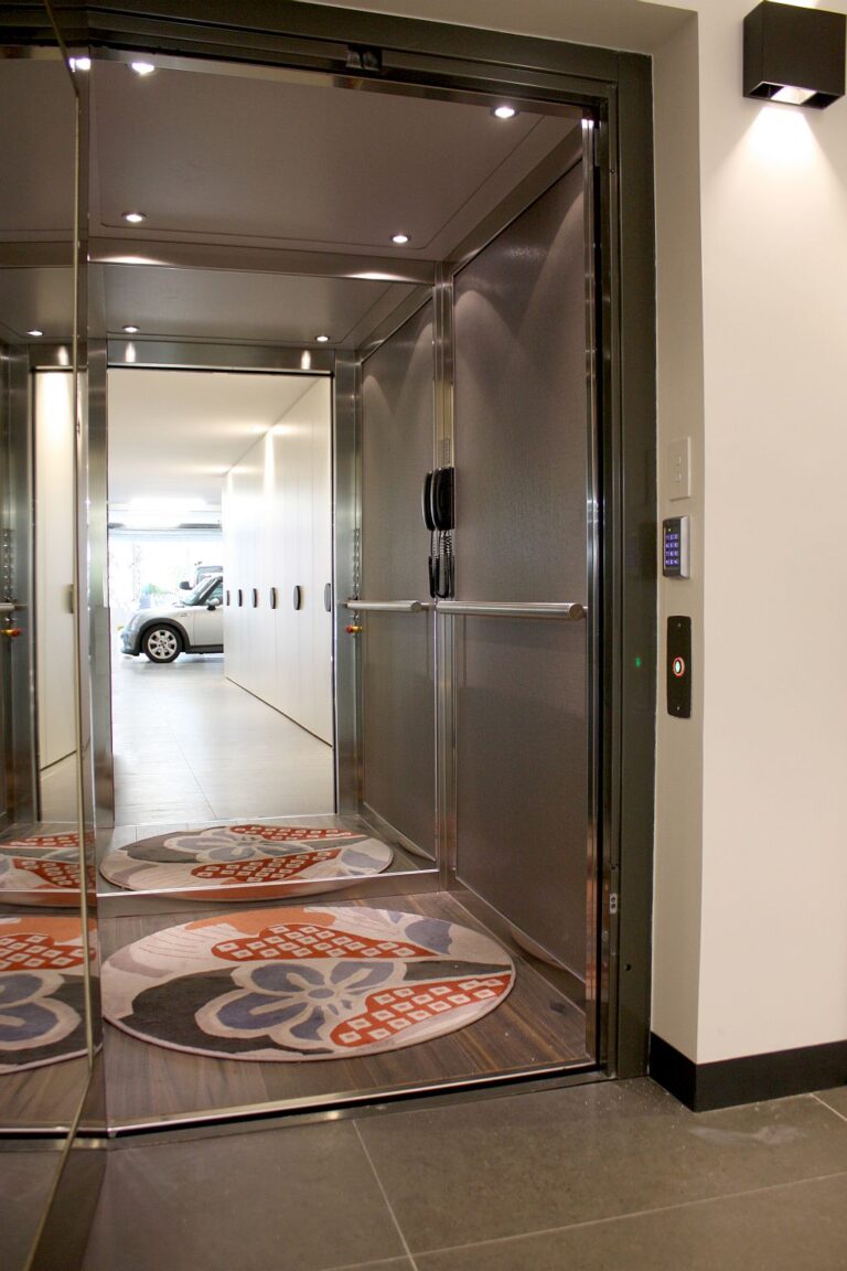 Lift cabin with stainless steel walls and a round patterned rug, looking through to the garage
