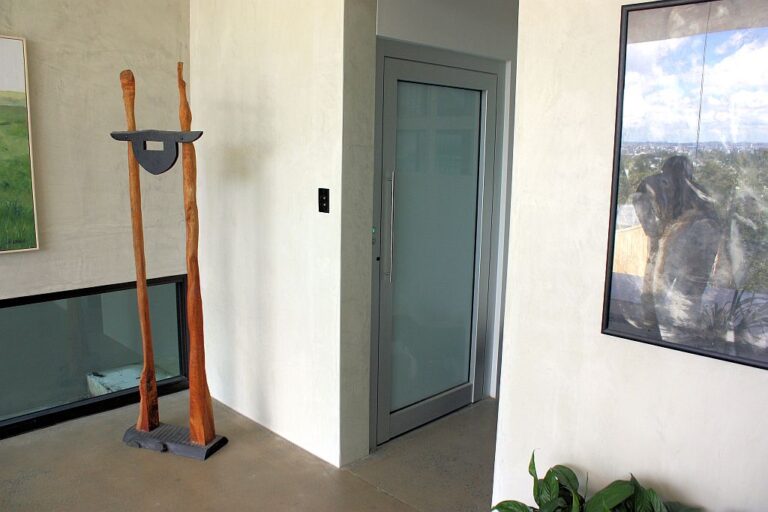 Hallway landing with the ElfoLift door in anodised aluminium and frosted glass, beside a timber sculpture and artwork