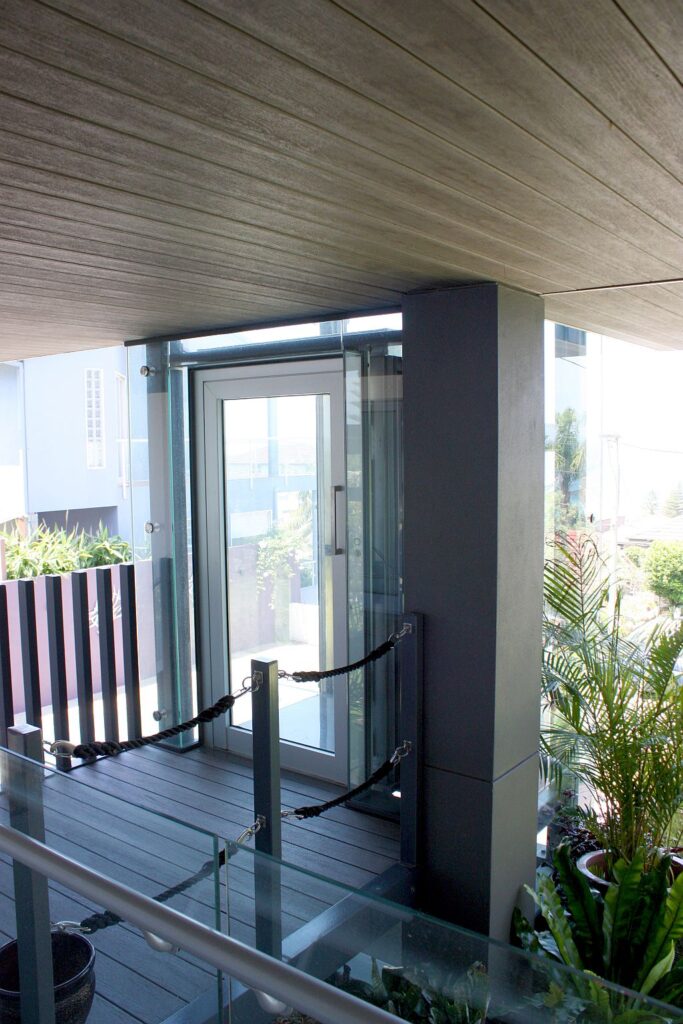 Anodised aluminium landing door with a transparent glass panel opening onto a deck beside a glass balustrade