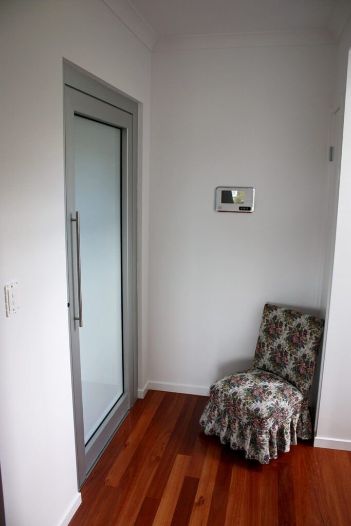 Lift landing door with a frosted glass panel in a white hallway beside a floral armchair