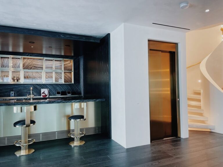 Bar with brass stools and a bronze finish lift door beside a curved white staircase