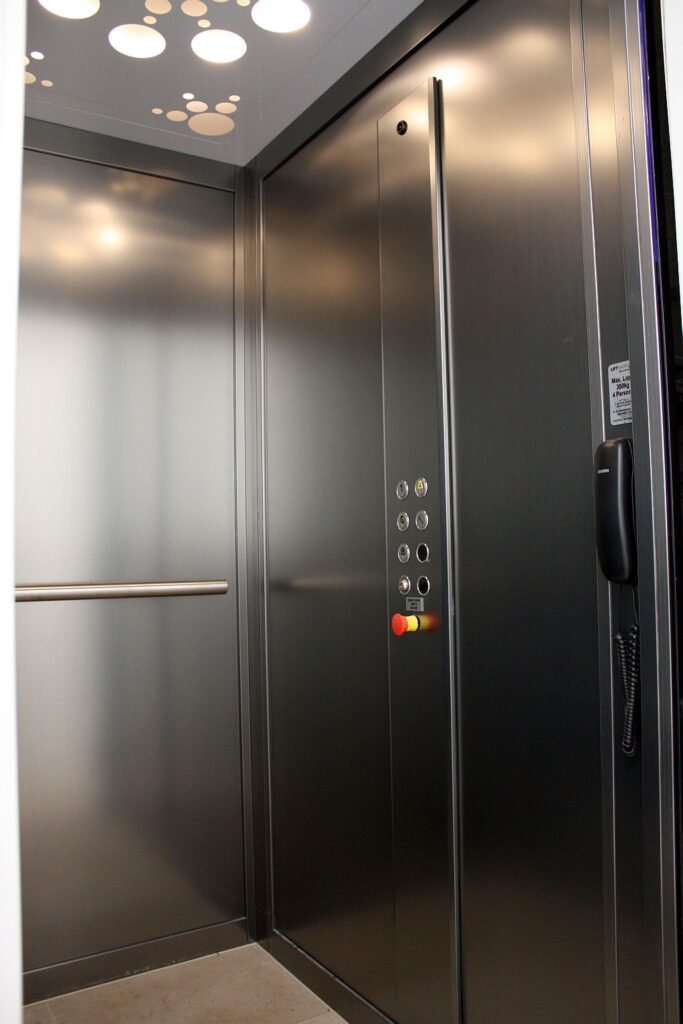 Inside a brushed stainless steel-look E1 cabin with a button panel, handrail and phone in Brighton