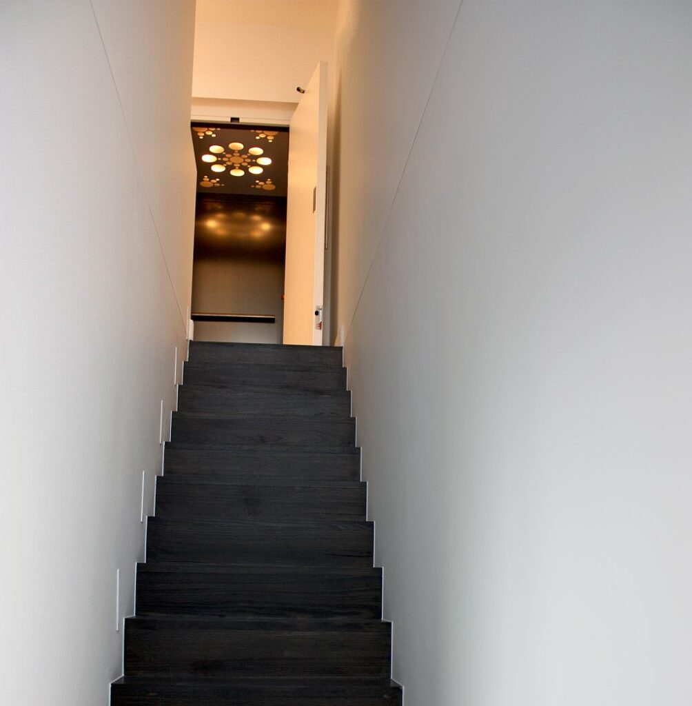 Dark staircase leading up to the lit E1 lift landing between white walls in a Brighton townhouse