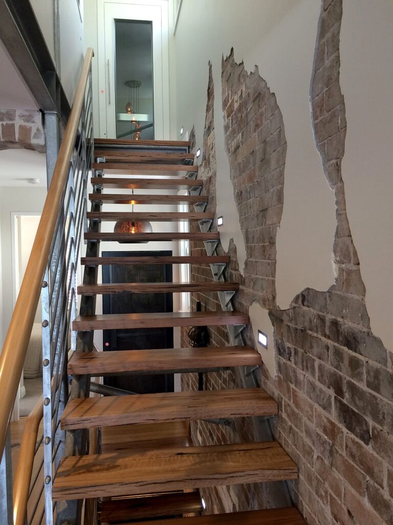 Timber staircase with exposed brick wall and stainless steel balustrade, with the Manly Cove home lift behind its glass door at the top