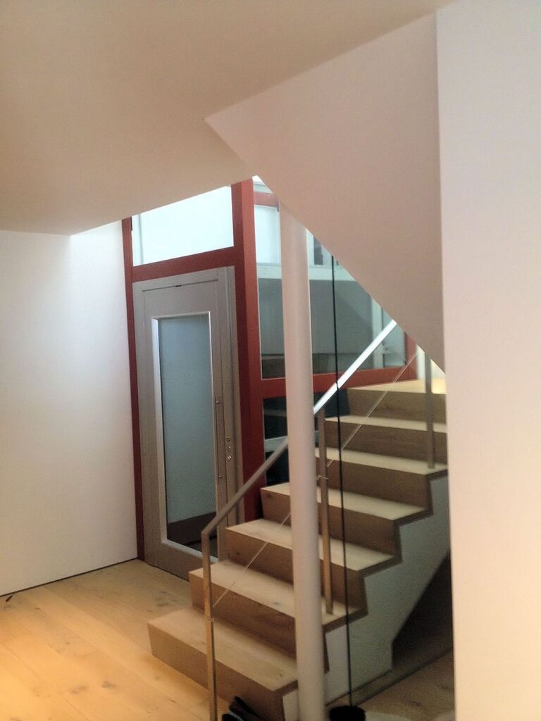 Timber stairs beside the red-framed glass lift shaft in a bright stairwell of the San Francisco home