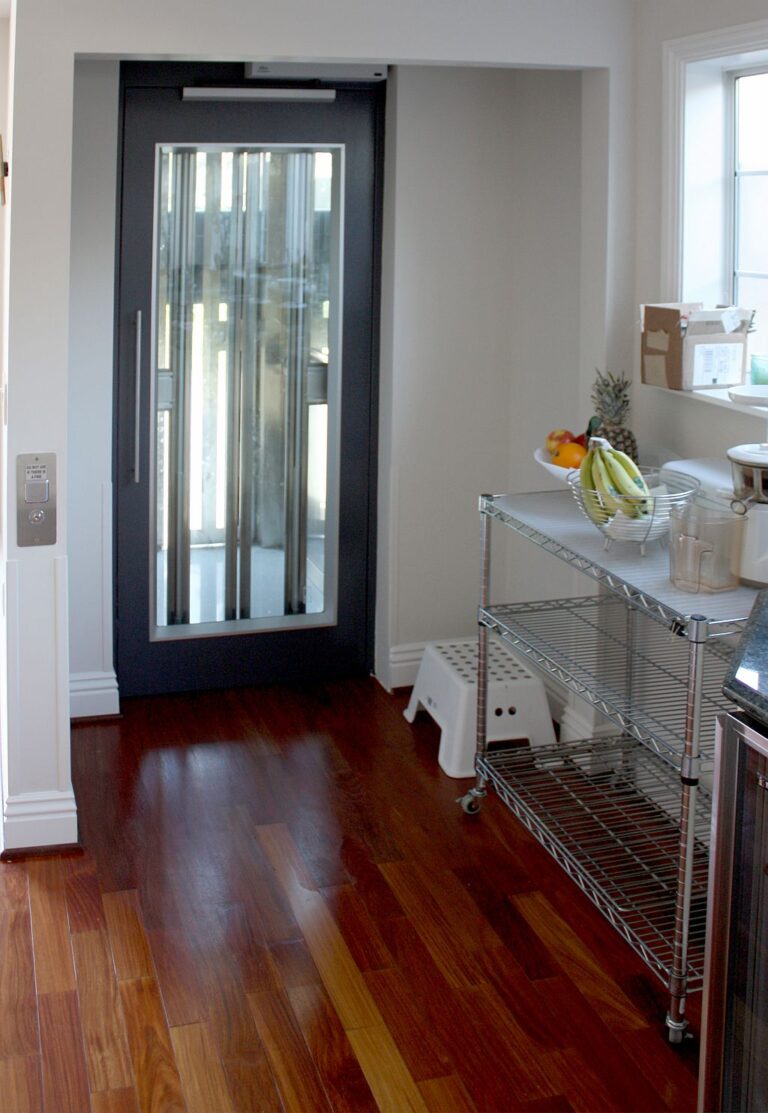 Glass-panelled lift door beside a kitchen shelf on a timber floor