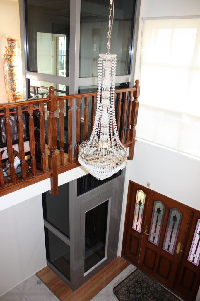 Looking down from the upper landing past a chandelier to the glass-shaft lift and the timber front door