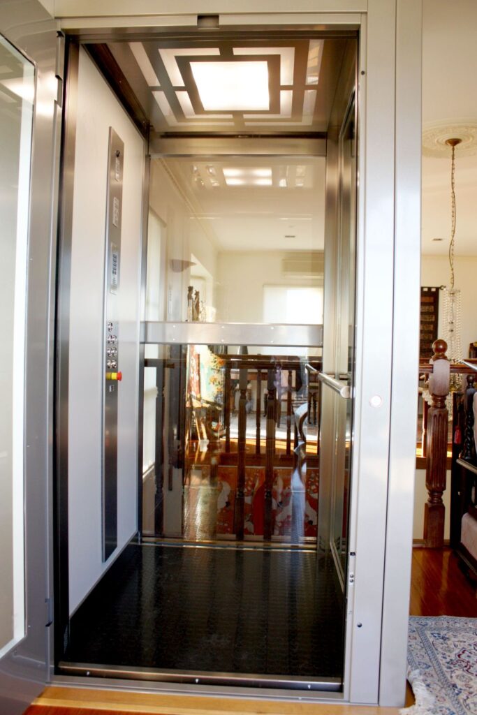 Inside the lift car with a glass door, steel control panel and a patterned ceiling light, looking through to the Hampton living area
