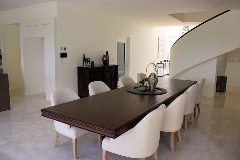 Open-plan dining room with a long dark timber table, cream chairs, marble-look tiles and the white lift door beside the curved staircase