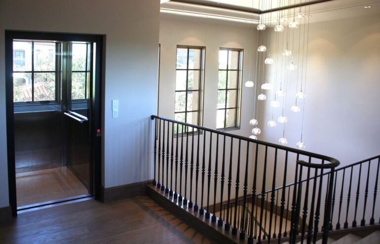 Open lift door on a landing beside a black balustrade and a cluster of pendant lights