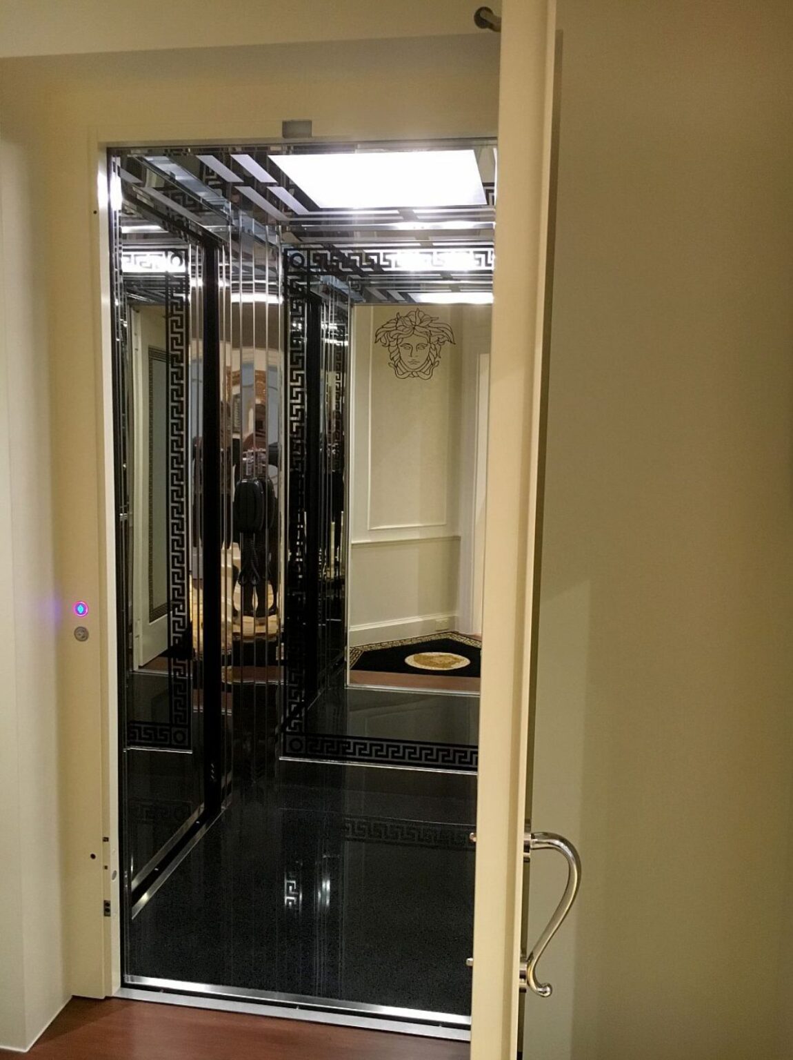 Open door of the E1 Classica home lift showing a black and mirrored cabin with a Versace Greek key border