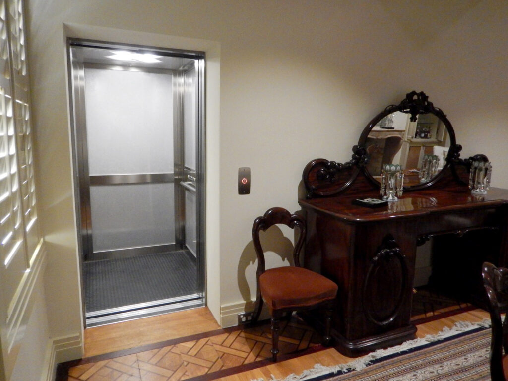 Open lift door showing the stainless steel cabin beside an antique sideboard and chair in a Melbourne home