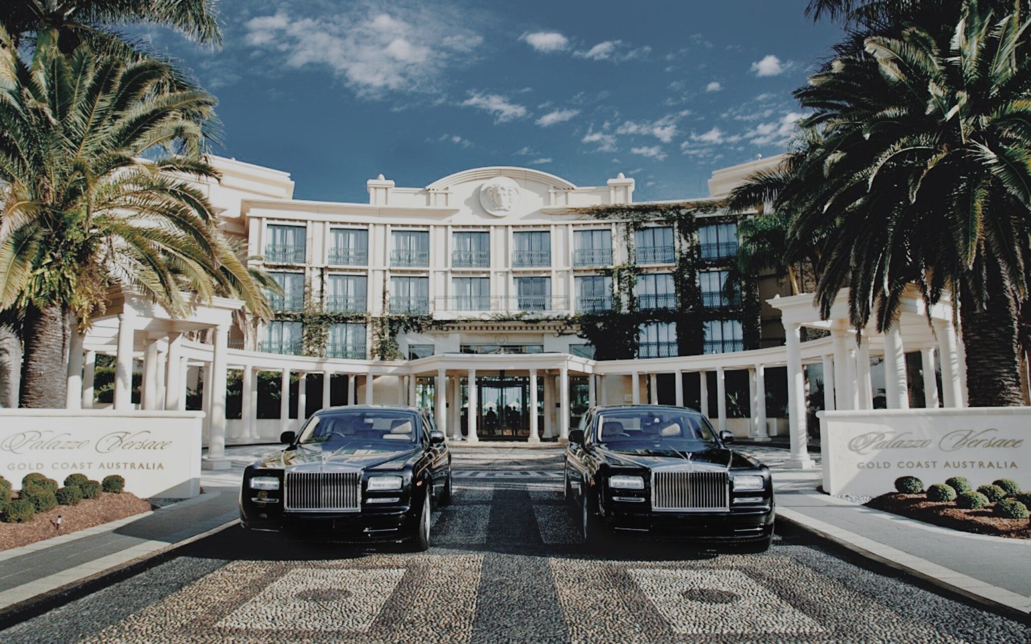 The Palazzo Versace entrance on the Gold Coast, with two black cars and palm trees in front of the cream building
