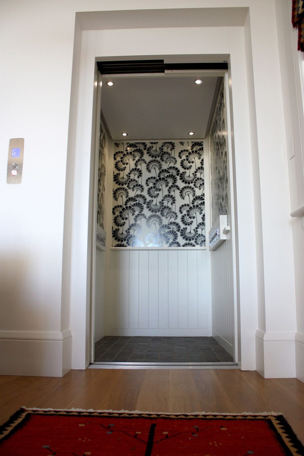 Open lift door showing a black and white floral wallpapered cabin, above a timber floor and red rug