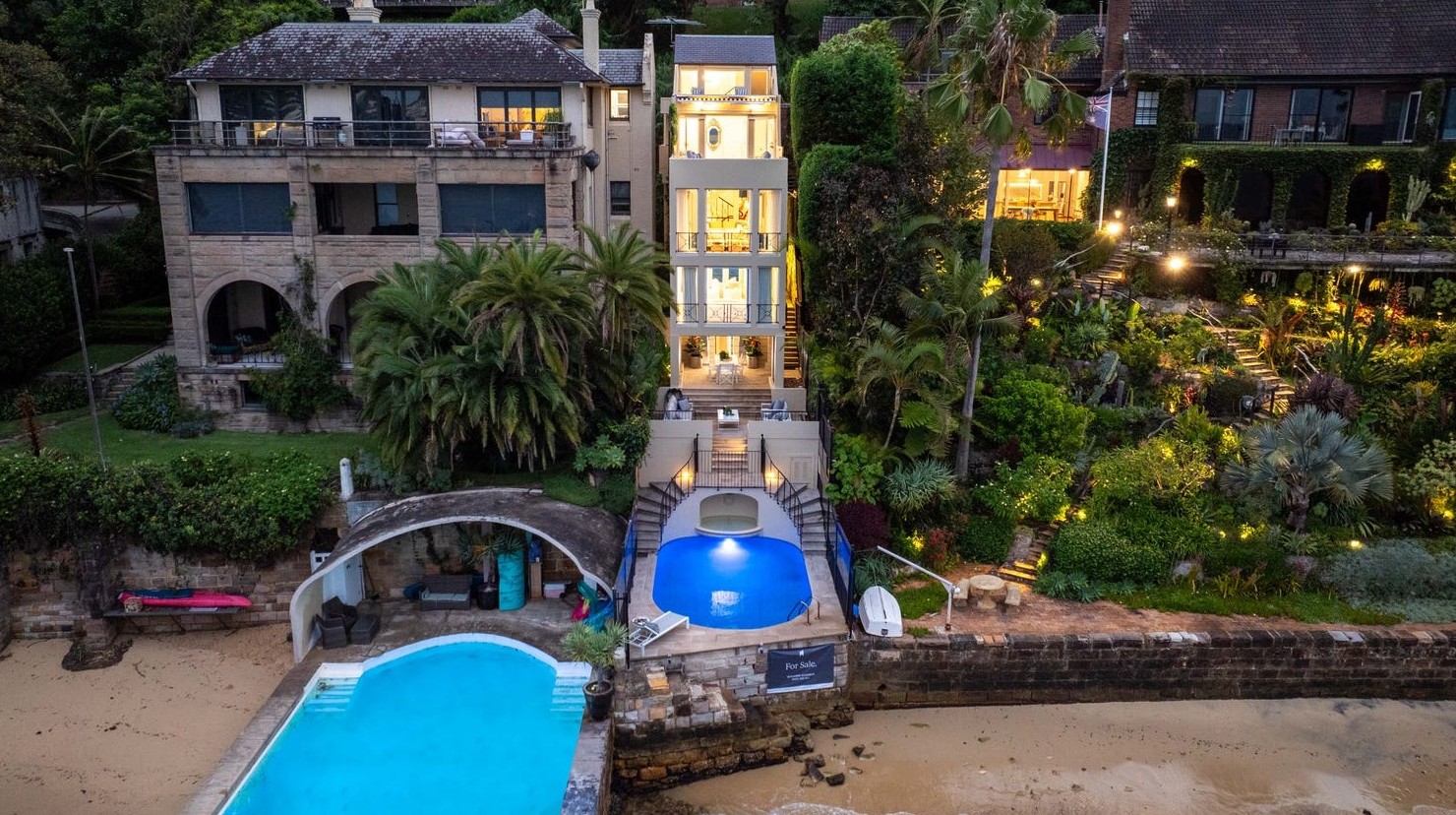 The narrow lit Darling Point home at dusk, rising above its pool and the sandy beach