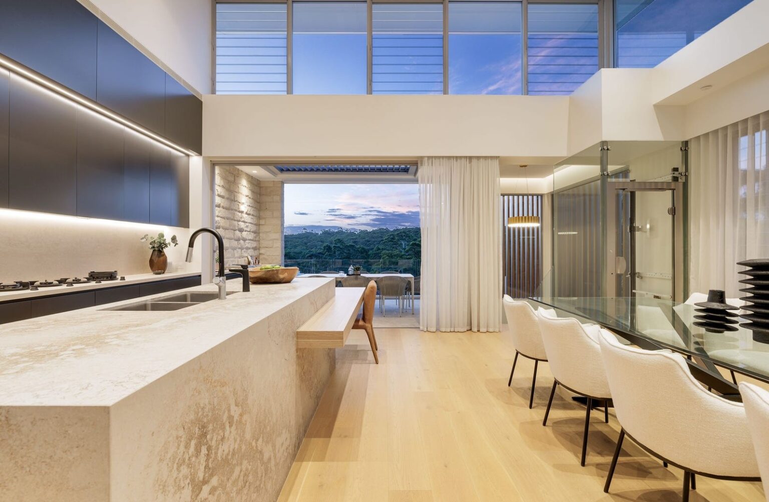 Kitchen island, dining table and terrace with a tall glass lift shaft at the right, Gladesville home