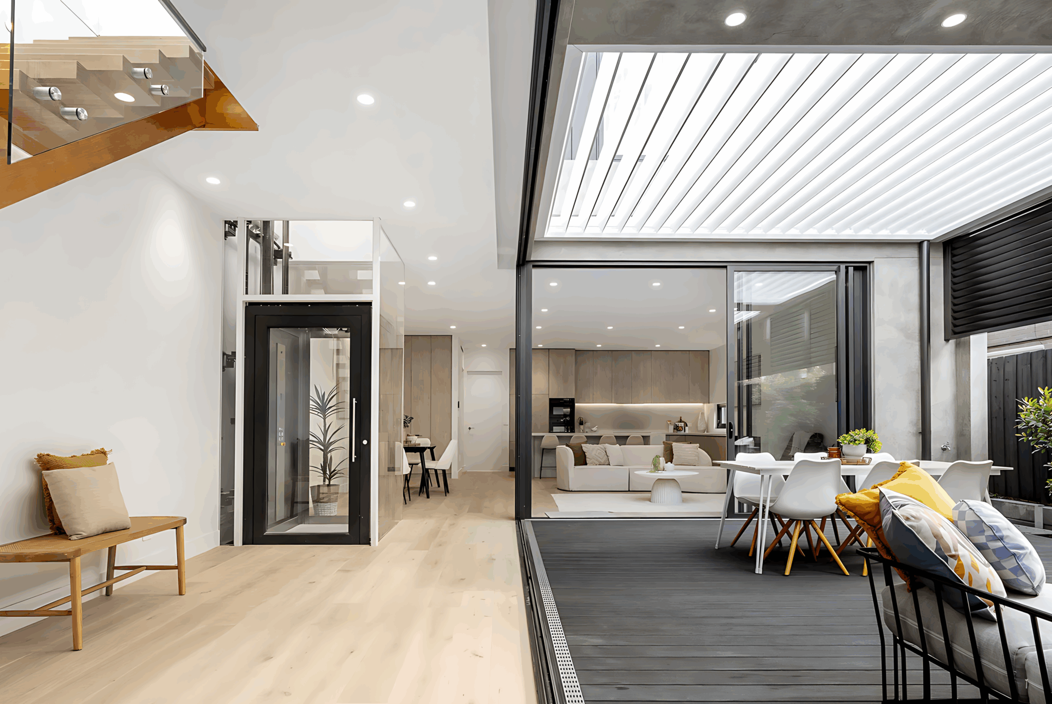 Glass-doored A Series lift with a matte black frame in the open-plan living space of the Caulfield North townhouse, beside a timber staircase and a louvred-roof deck