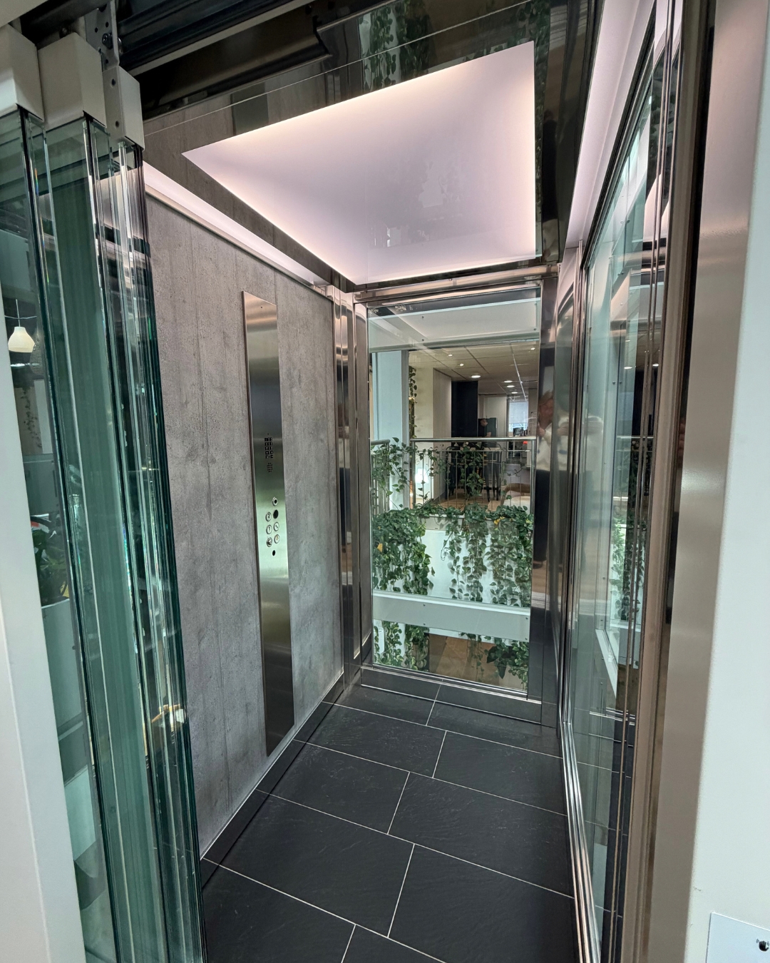 Inside a home lift cabin with glass walls, stainless steel trims, a lit ceiling and a dark tiled floor