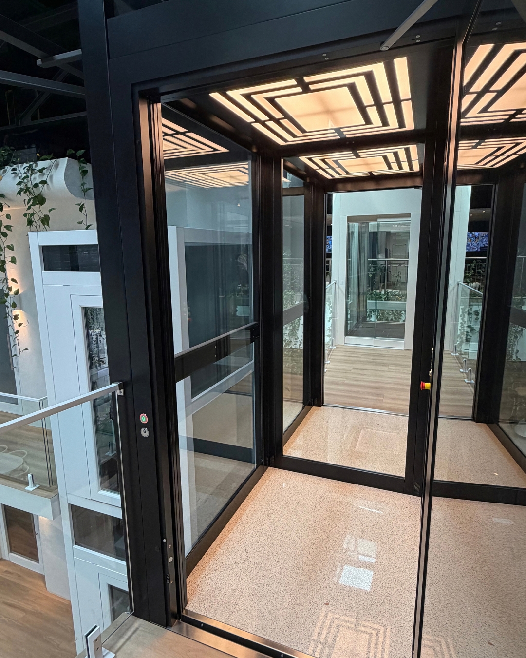 Home lift cabin with full-height glass walls in a black frame, under a patterned lit ceiling