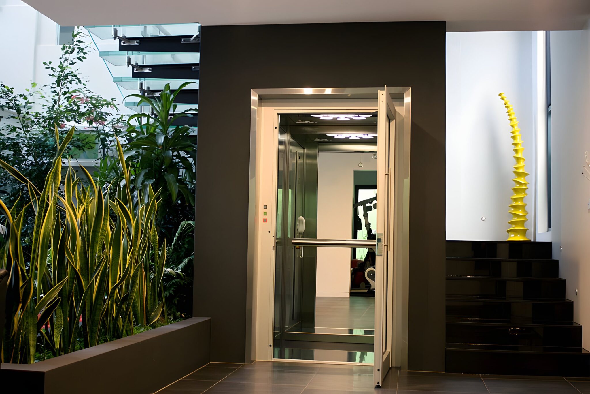 Home lift with a glass-panelled door and stainless steel surround set into a dark feature wall, beside a black stone staircase and indoor plants in a Canberra home
