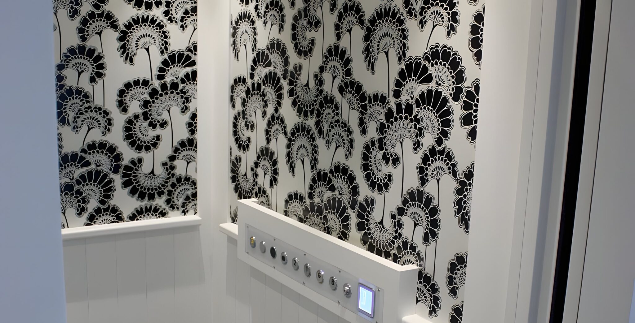 Lift cabin wall in black and white floral wallpaper above white panelling, with a row of push buttons