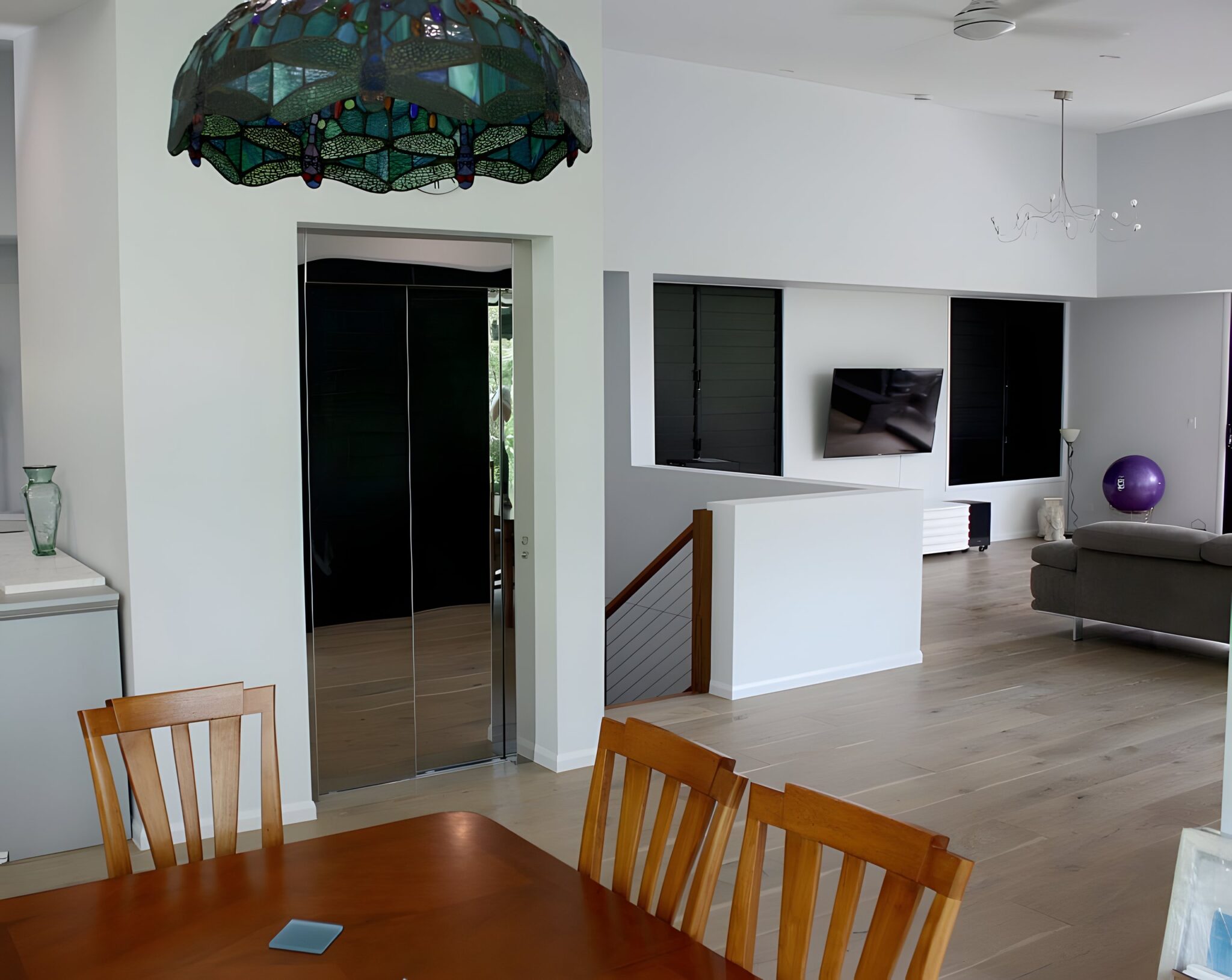 Black and stainless steel home lift doors beside a dining table and stained glass lamp in a Queensland home