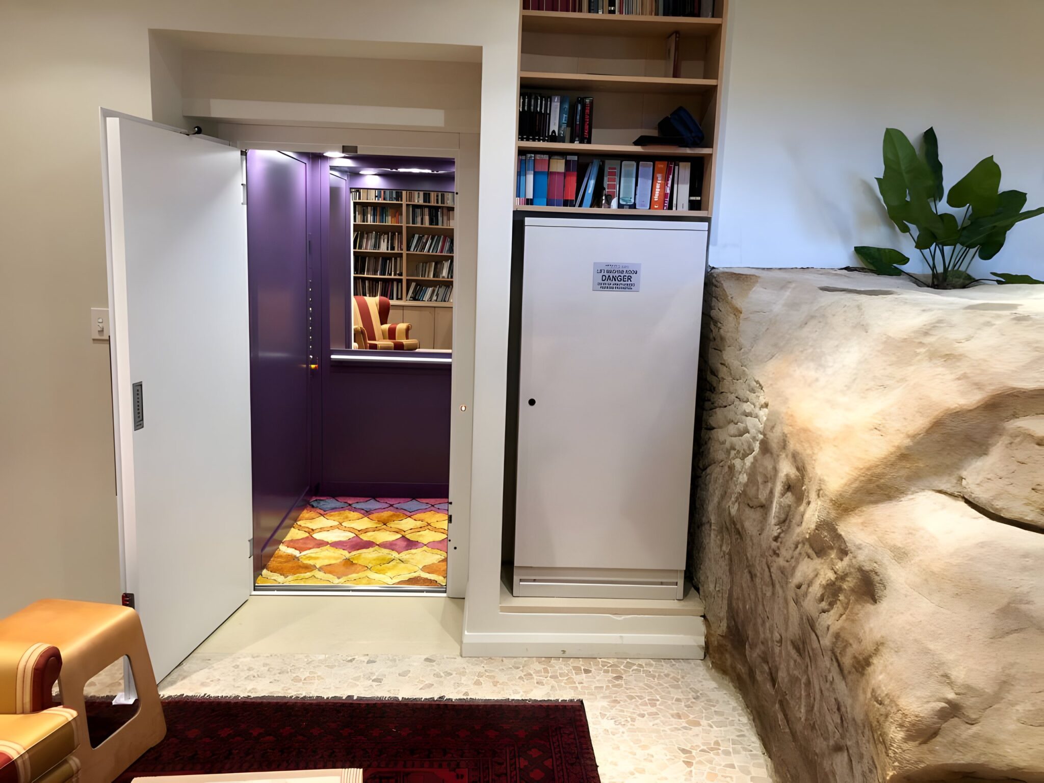 Open lift door showing a purple cabin with a patterned floor beside a bookshelf and a sandstone look rock feature