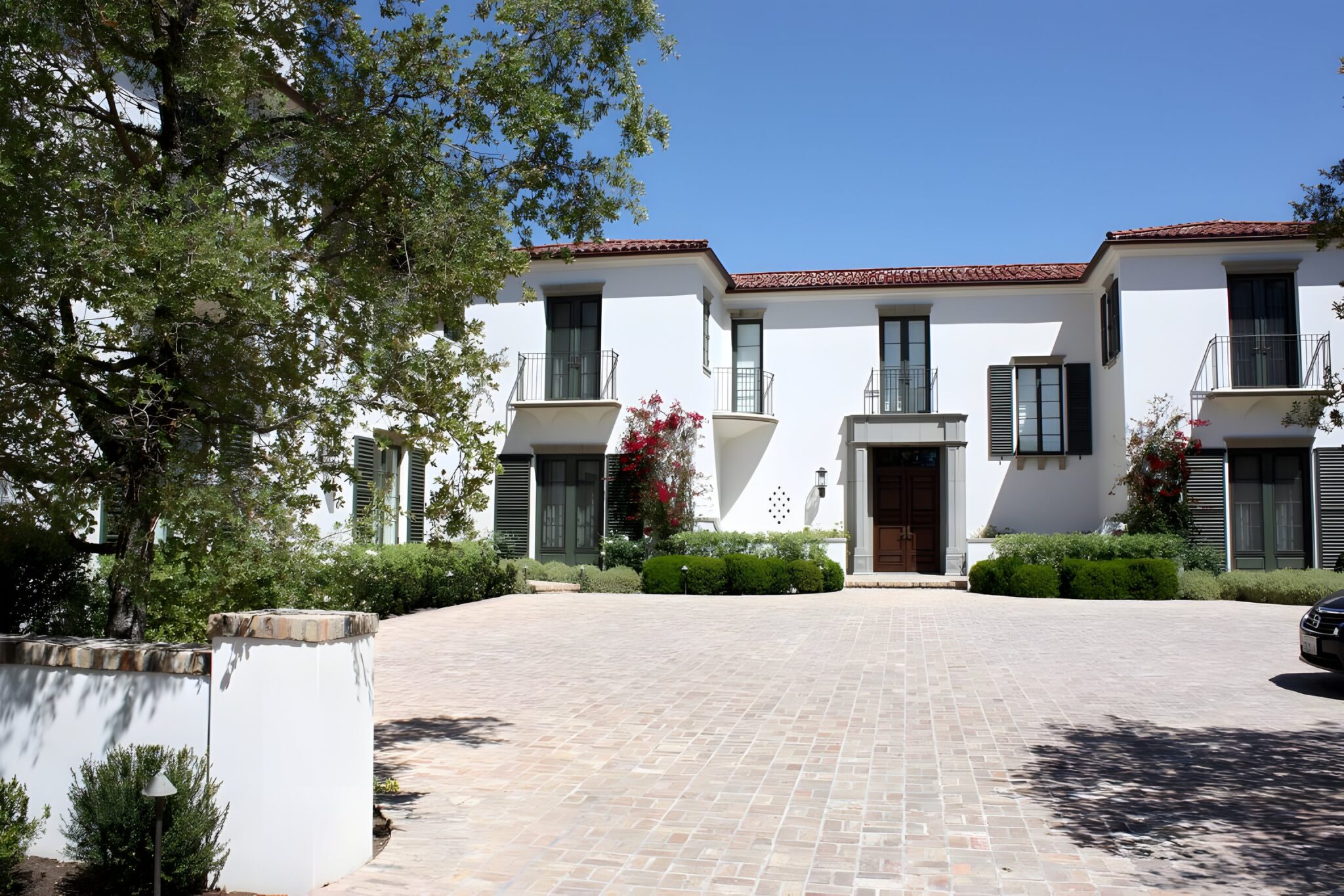 Two-storey white Spanish-style home with a terracotta tile roof, green shutters and a paved driveway in Santa Monica, California