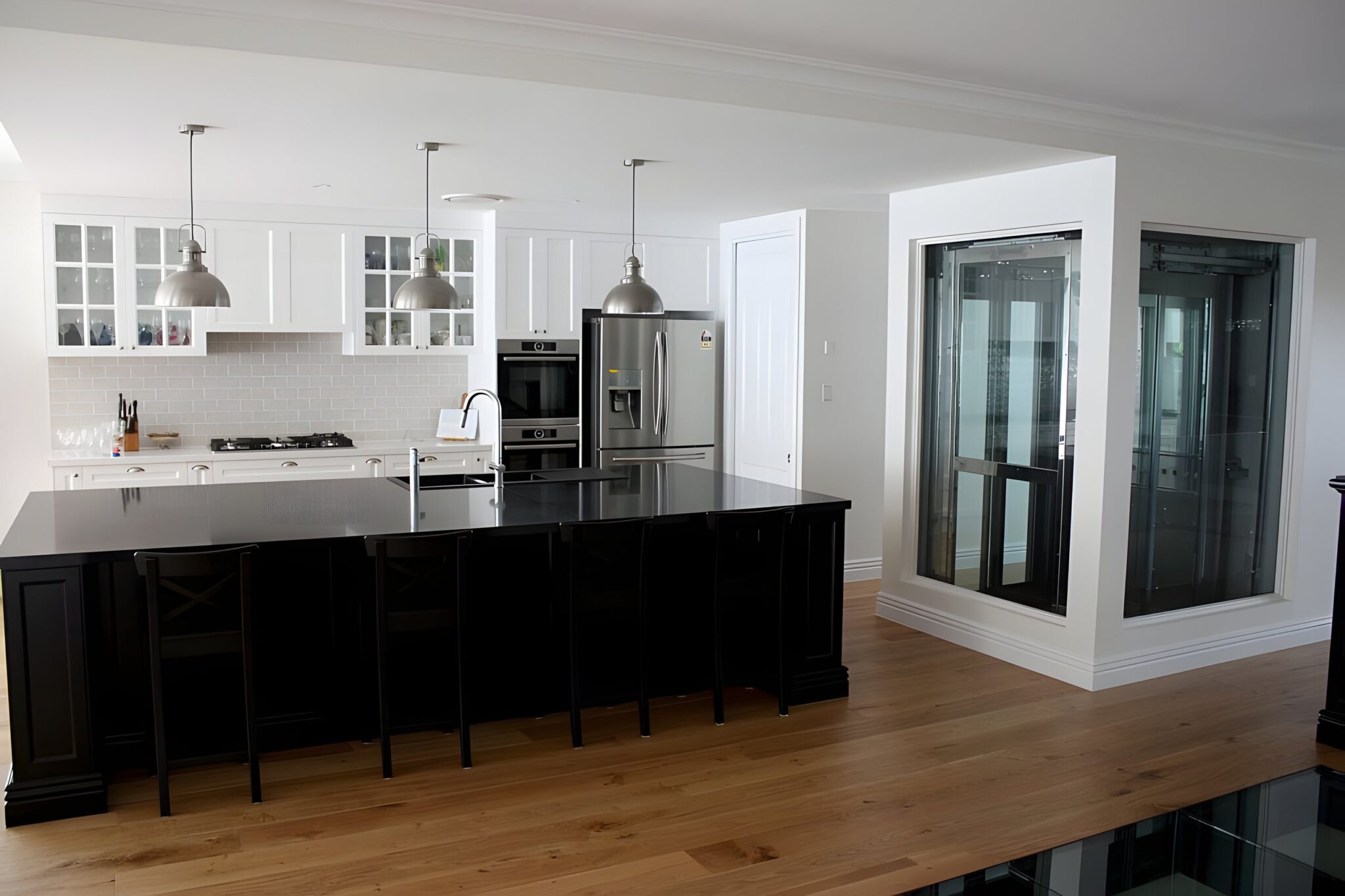 Open-plan kitchen with a black island and a glass lift shaft in a white surround at the back of the room