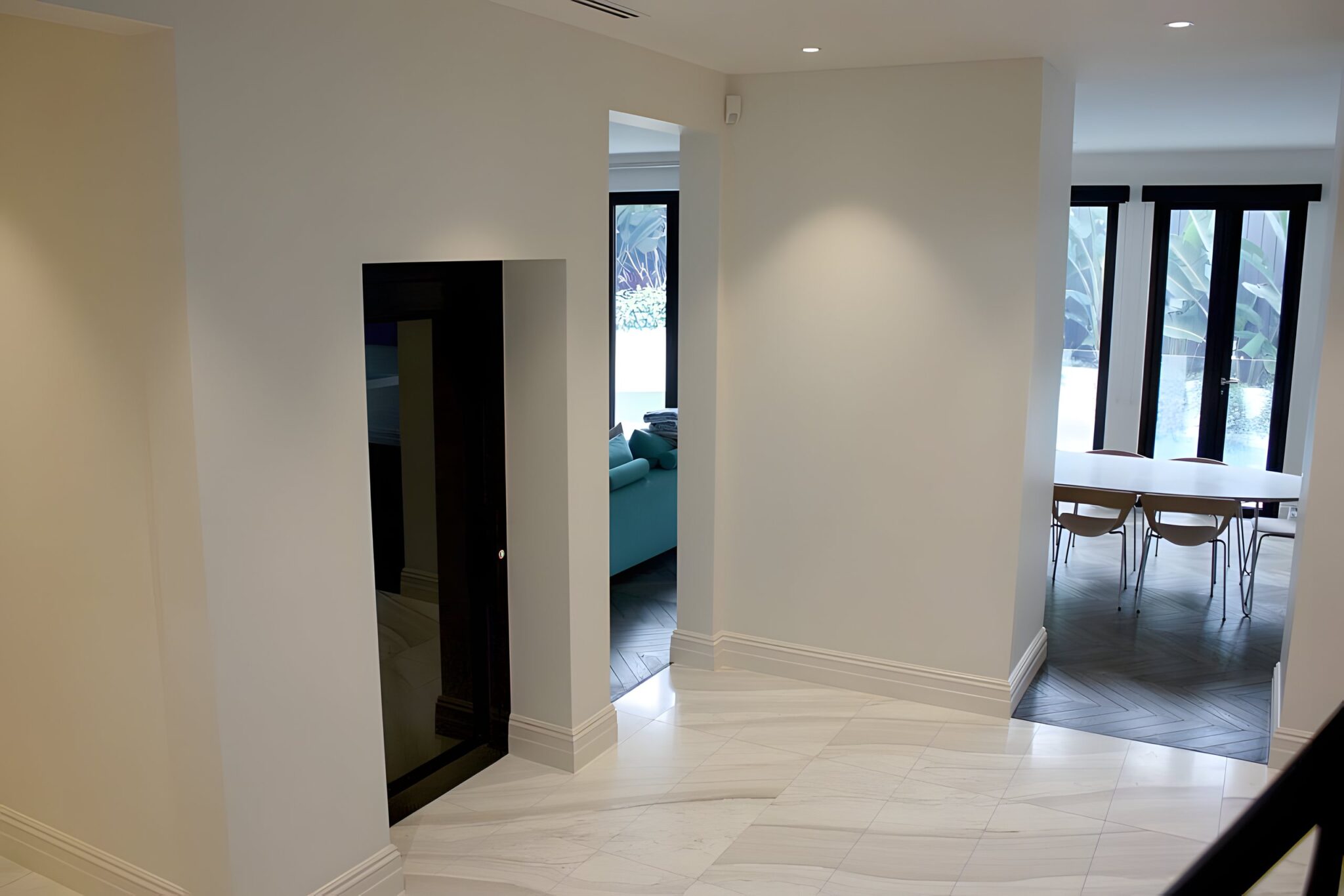 Black glass lift door in a white corridor leading to a dining area with glass doors in the Melbourne home