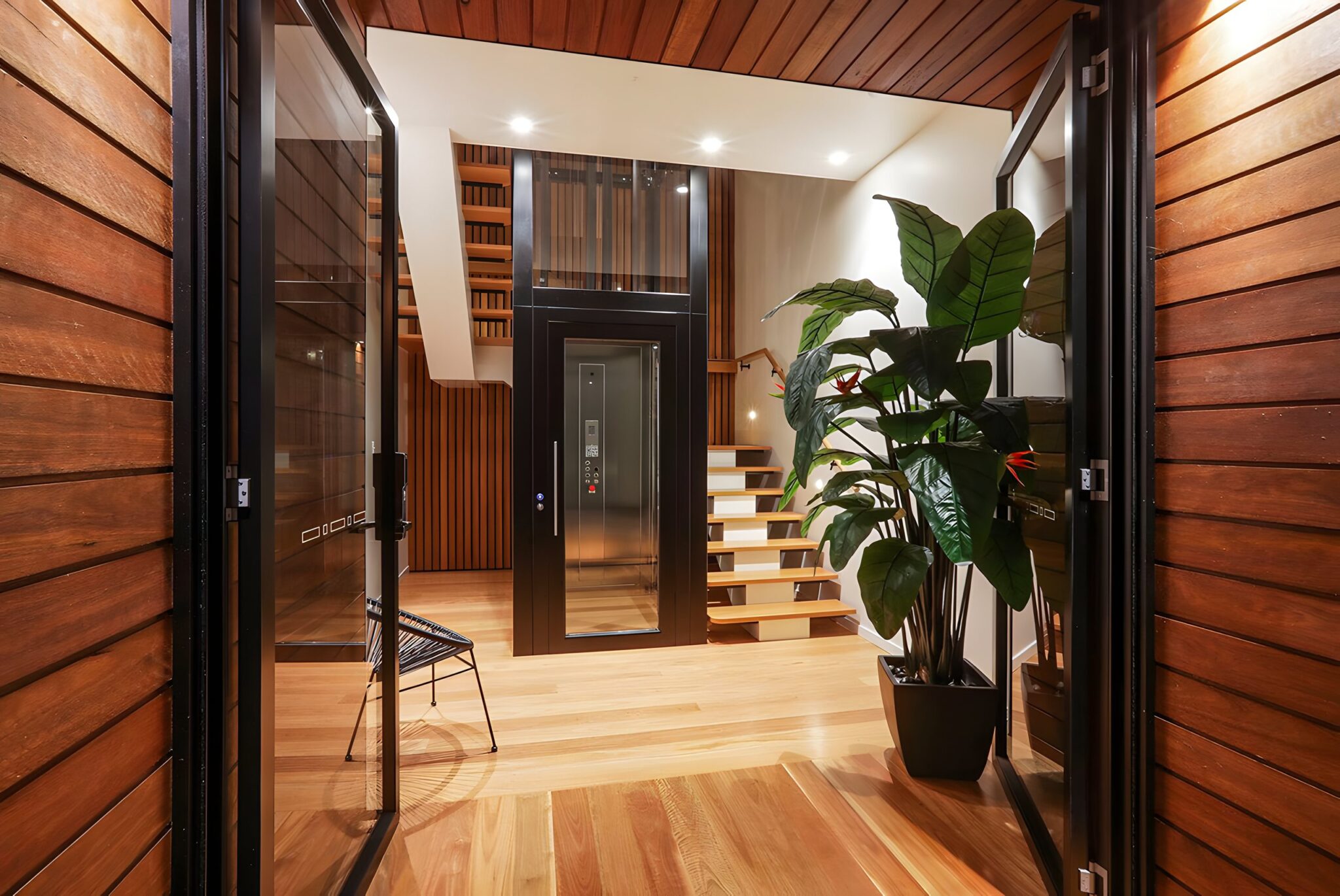 Black framed glass home lift seen through timber clad doors, beside open timber stairs and a large indoor plant in a Currumbin home