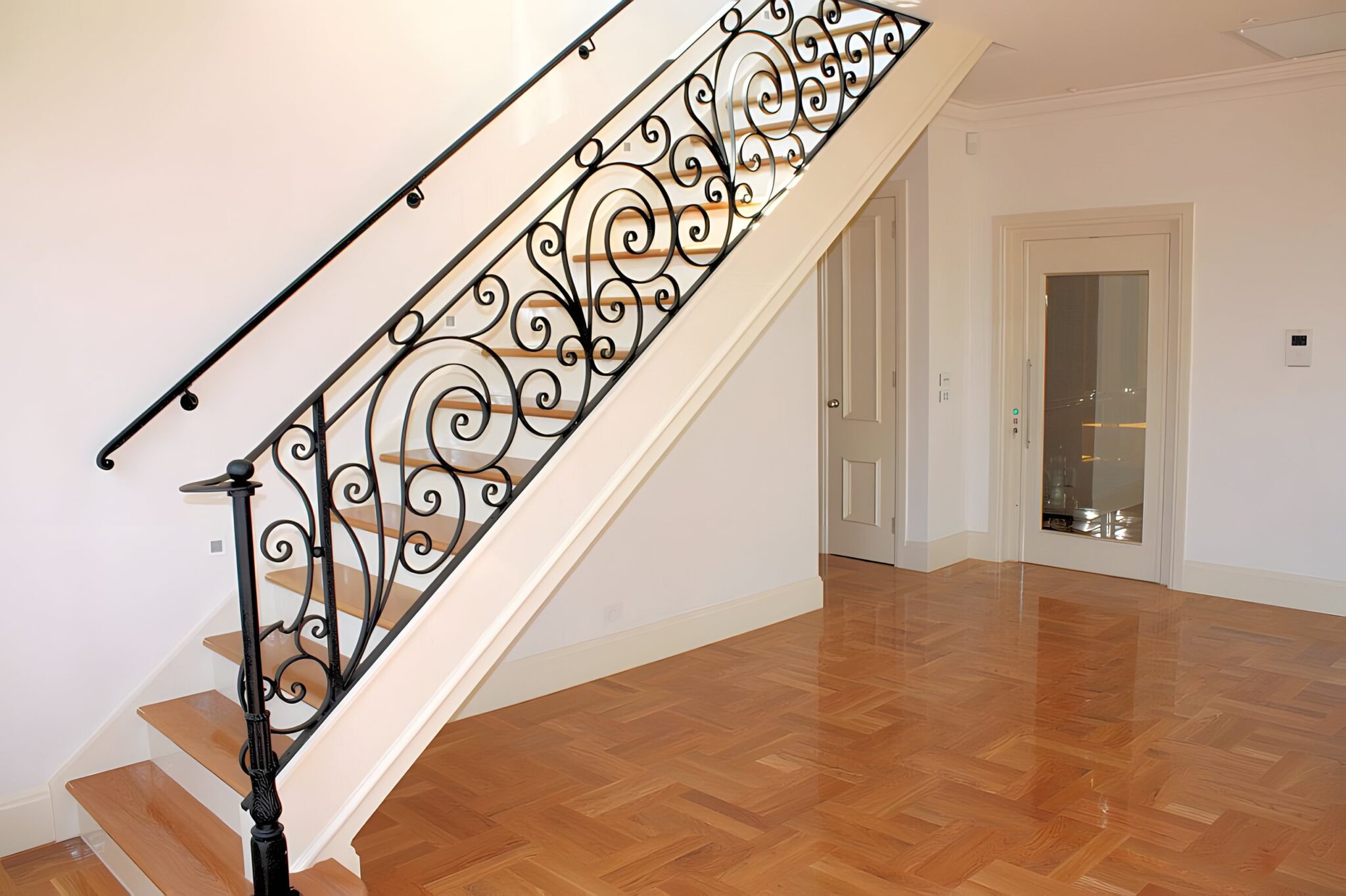 Timber staircase with a wrought iron balustrade above a polished parquet floor, the glass-fronted lift door beside it in a Melbourne home