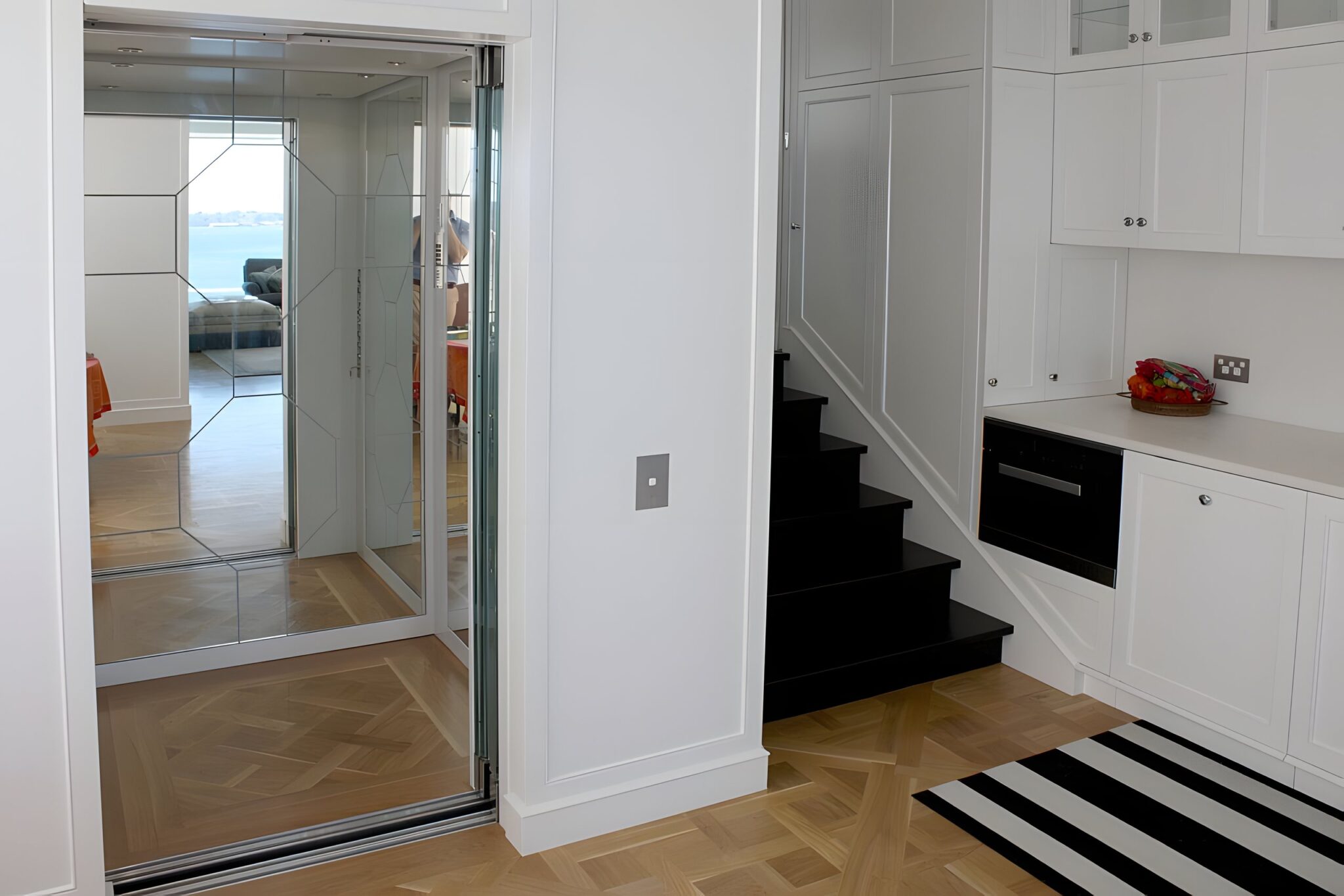 Open glass lift door with geometric mirror panels inside, beside a black staircase and a striped rug on a parquetry floor