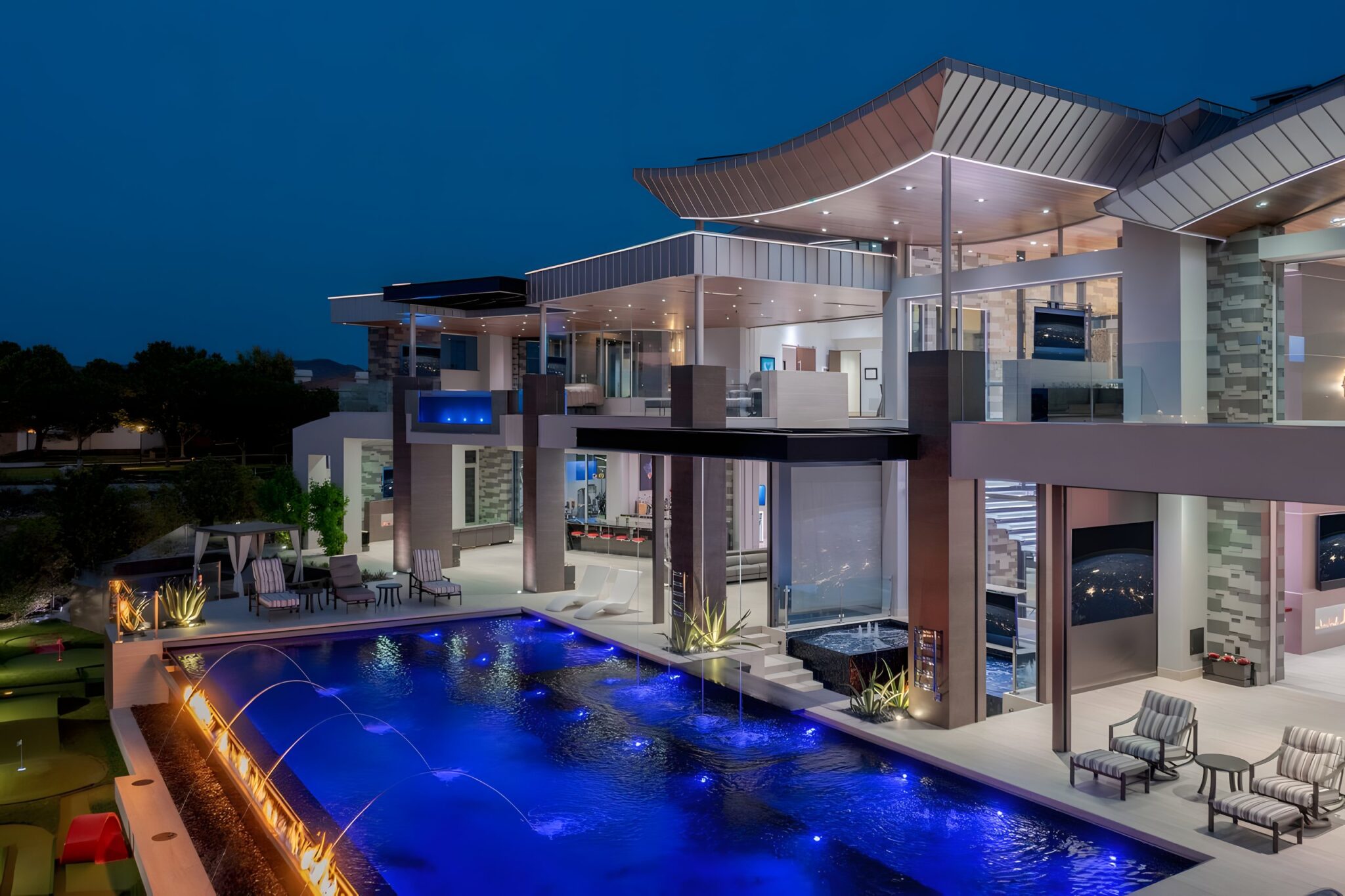 Modern Henderson, Nevada home at night with a lit swimming pool, fire feature, terraces and glass walls under curved timber-soffit roofs