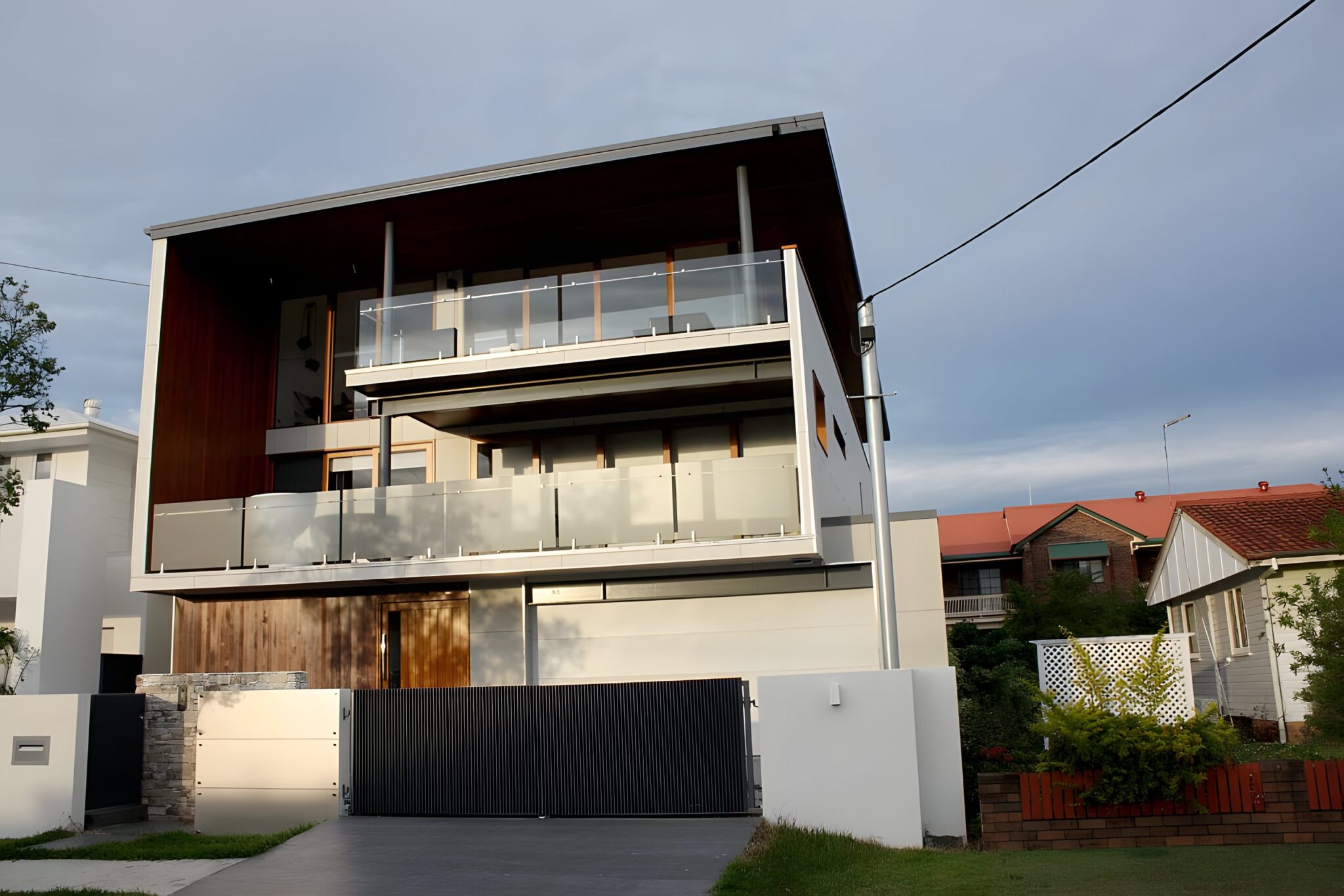 Modern three-storey home in Bulimba, Brisbane, with timber cladding, a timber front door and glass balustrades at dusk